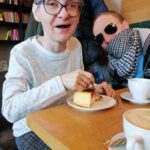 Starsza kobieta w okularach siedząca przy stoliku pijąca kawę i jedząca ciastko. W tle mężczyzna z zasłoniętymi ustami chustą w drobną biało-czarną szachownicę.